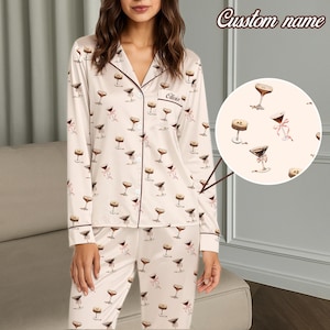 May include: Cream-coloured pyjama set featuring a pattern of martini glasses and pink bows. The long-sleeved top and trousers have dark brown trim. The top has a breast pocket with the name "Olivia" embroidered. The words "Custom name" are at the top.