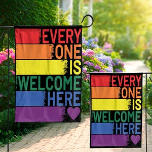 Może przedstawiać: Dwie flagi ogrodowe z tęczowym wzorem i napisem "EVERYONE IS WELCOME HERE". Flagi mają czarne tło z poziomymi paskami w kolorach czerwonym, pomarańczowym, żółtym, zielonym, niebieskim i fioletowym. Na dole fioletowe serce.