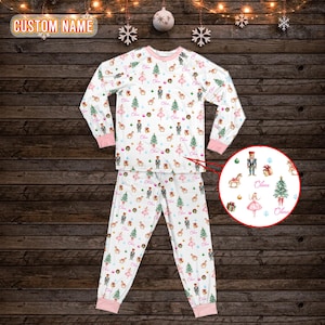 May include: White pajama set with a repeating pattern of Christmas trees, ballerinas, nutcrackers, and gift boxes. The set has pink trim and the name "Olivia" is printed on the design. The pajamas are displayed on a dark wood background.