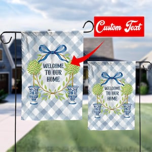 Könnte beinhalten: Eine blaue und weiße karierte Gartenflagge mit einem Kranz aus grünen Blättern und einem blauen Band. Der Kranz hat zwei Formschnittbäume in blauen und weißen Urnen. Der Text "Willkommen in unserem Zuhause" befindet sich in der Mitte des Kranzes.