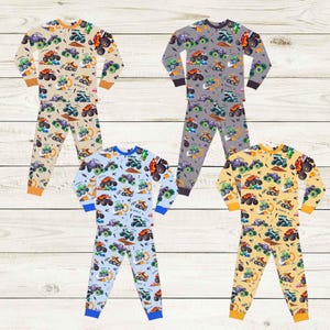May include: Four sets of children's pajamas with a repeating monster truck pattern. The pajamas are in beige, gray, light blue, and yellow. Each set includes a long-sleeved top and pants. The cuffs and necklines are in contrasting colors.