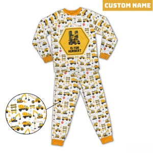Può includere: Pigiama bianco con un motivo ripetuto di veicoli da costruzione gialli. La maglia a maniche lunghe presenta un grande esagono giallo con un veicolo da costruzione e la scritta "I IS FOR HUBERT". Il pigiama ha finiture arancioni.