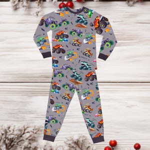 May include: Gray pajama set with a repeating pattern of colorful monster trucks. The long-sleeved top and pants feature various trucks in green, purple, orange, and blue, with the words "WOOOM" and "CRUSH IT!" scattered throughout.