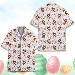 May include: White short-sleeved button-down shirt featuring a repeating pattern of a brown Bigfoot carrying a basket and a white unicorn with an Easter egg basket. The shirt has a collared neckline and is displayed flat and folded.