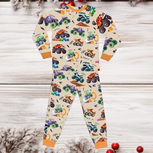 May include: Beige pajama set with a repeating pattern of colorful monster trucks. The long-sleeved top and pants feature orange trim and the words "VROOOM" and "CRUSH IT!" scattered throughout the design. The pajamas are made of a soft material.