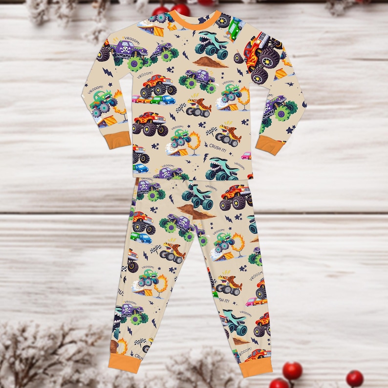May include: Beige pajama set with a repeating pattern of colorful monster trucks. The long-sleeved top and pants feature orange trim and the words "VROOOM" and "CRUSH IT!" scattered throughout the design. The pajamas are made of a soft material.