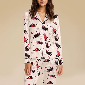 Puede incluir: Conjunto de pijama color crema con un estampado de gatos negros y corazones. La parte superior de manga larga tiene cuello de solapa y ribete negro. Los pantalones a juego completan el conjunto. Los gatos llevan sombreros de fiesta y vestidos.