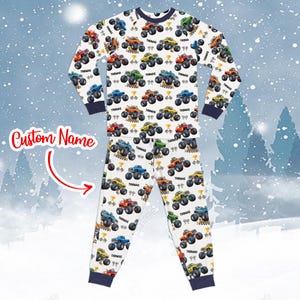 Peut inclure: Ensemble de pyjama une pièce blanc avec un motif répété de monster trucks colorés. Chaque camion porte le nom "THOMAS". Le pyjama a des bordures bleu marine sur le col, les poignets et les chevilles. Le texte "Custom Name" est également visible.