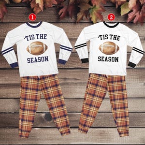 Peut inclure: Deux ensembles de pyjamas assortis. Chaque ensemble comprend un haut blanc à manches longues avec un motif de ballon de football et le texte "'Tis the Season". Les pantalons à carreaux sont marron, orange et bleu. Un ensemble a des accents bleu marine, l'autre noir.