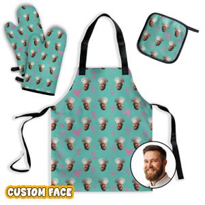 May include: A kitchen set featuring an apron, two oven mitts, and a square pot holder, all in a teal color with a repeating pattern of a man's face wearing a chef's hat and pink heart outlines. The apron includes black straps and ties.