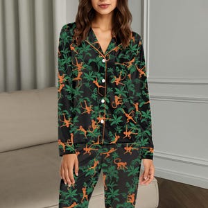 May include: A black pajama set featuring a long-sleeved button-down shirt and matching pants. The fabric displays a repeating pattern of orange monkeys and green tropical leaves. The shirt includes a notched collar and orange piping trim.