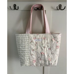 May include: A quilted tote bag featuring a patchwork design with a cream-colored fabric adorned with pink roses and a sage green plaid pattern. The bag has two light pink fabric handles and a small white rectangular label with text.