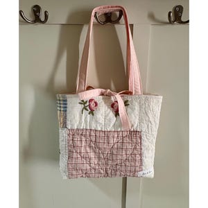 May include: A quilted tote bag featuring a patchwork design with cream, blue, and red plaid patterns. The front displays two embroidered pink roses with green leaves and a tied pink fabric bow at the top. A small white label reads Love & Gin.