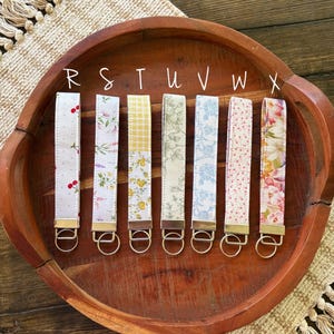 May include: Seven fabric key fobs with different patterns, including cherries, floral, and gingham, are displayed in a wooden bowl. Each fob has a gold-colored metal ring. The letters R, S, T, U, V, W, and X are written above each fob.