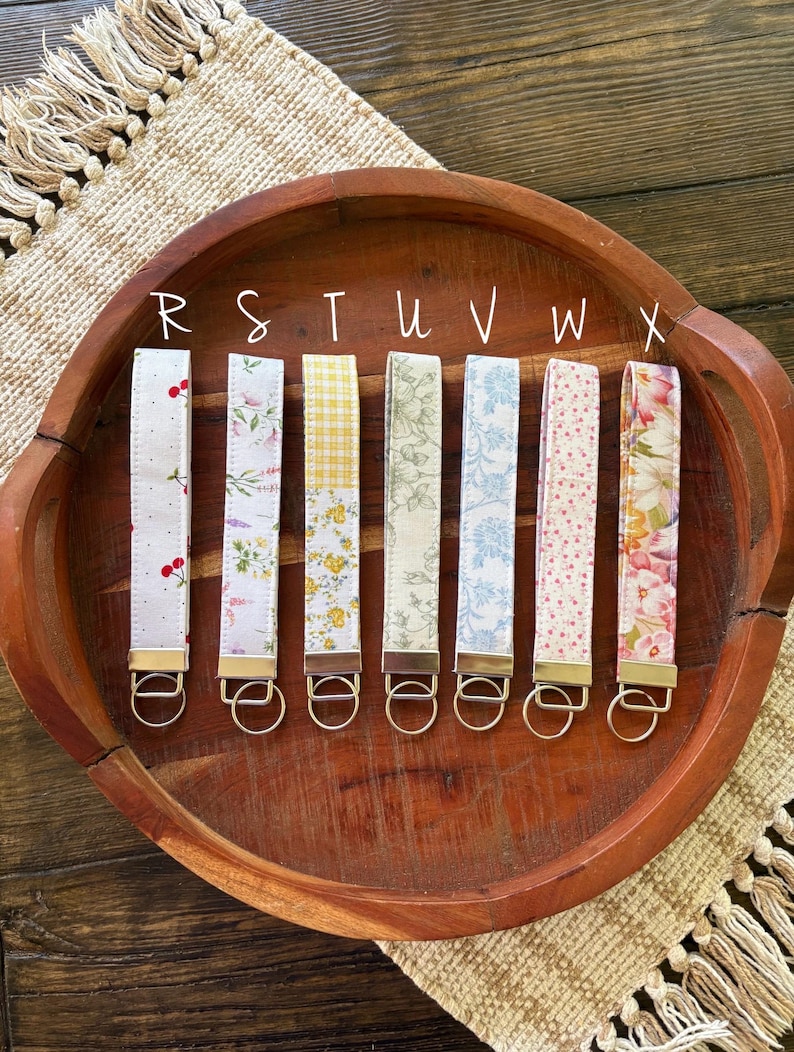 May include: Seven fabric key fobs with different patterns, including cherries, floral, and gingham, are displayed in a wooden bowl. Each fob has a gold-colored metal ring. The letters R, S, T, U, V, W, and X are written above each fob.