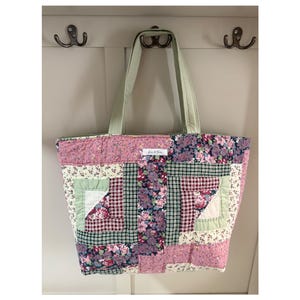 May include: A quilted tote bag featuring a patchwork design of floral and checkered fabrics in shades of pink, green, and white. The bag has two light green fabric handles and a small white rectangular label with text centered at the top.