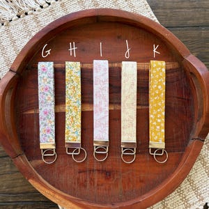 May include: Five floral fabric wrist keychains in various colors, including pink, yellow, and blue, with gold-colored hardware. The keychains are arranged on a wooden tray with a woven placemat.