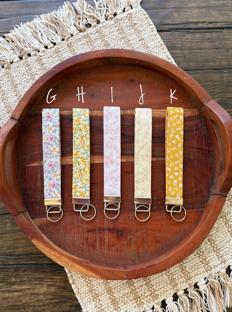 May include: Five floral fabric wrist keychains in various colors, including pink, yellow, and blue, with gold-colored hardware. The keychains are arranged on a wooden tray with a woven placemat.