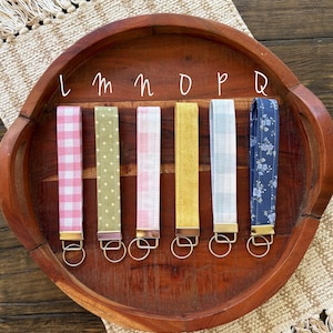 May include: A collection of six key fobs with various fabric patterns, including pink gingham, green polka dots, and floral designs. Each fob has a gold-colored metal clasp and a key ring. The fobs are arranged on a wooden tray.