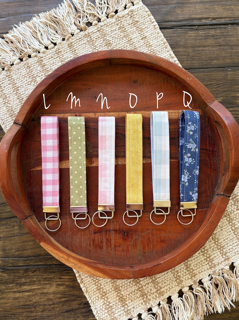 May include: A collection of six key fobs with various fabric patterns, including pink gingham, green polka dots, and floral designs. Each fob has a gold-colored metal clasp and a key ring. The fobs are arranged on a wooden tray.