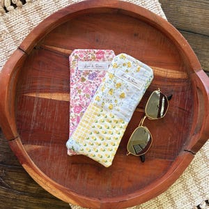 Puede incluir: Dos estuches para gafas con estampado floral y unas gafas de sol sobre una bandeja de madera. Un estuche es rosa con un estampado floral, el otro es amarillo con un estampado floral. Las gafas de sol tienen montura dorada y lentes oscuras. Los estuches tienen el texto "Love & Rose".