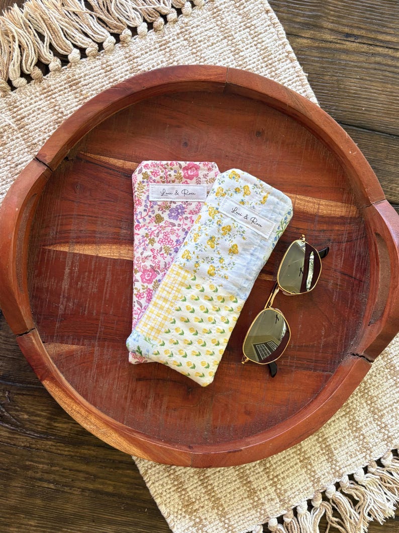 Puede incluir: Dos estuches para gafas con estampado floral y unas gafas de sol sobre una bandeja de madera. Un estuche es rosa con un estampado floral, el otro es amarillo con un estampado floral. Las gafas de sol tienen montura dorada y lentes oscuras. Los estuches tienen el texto "Love & Rose".