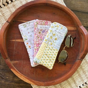 Puede incluir: Tres estuches acolchados para gafas con estampados florales en lavanda, rosa y amarillo, junto con un par de gafas de sol. Los estuches llevan la etiqueta "Love & Rose" y descansan sobre una bandeja de madera.