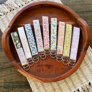 May include: A collection of key fobs with various floral patterns, including blue, pink, and yellow, displayed in a wooden tray. Each fob has a gold-colored metal ring and a leather-like backing. The tray sits on a beige woven placemat.