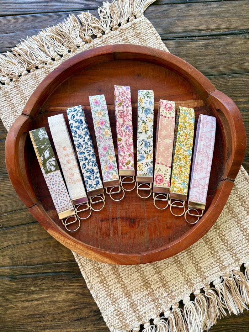 May include: A collection of key fobs with various floral patterns, including blue, pink, and yellow, displayed in a wooden tray. Each fob has a gold-colored metal ring and a leather-like backing. The tray sits on a beige woven placemat.