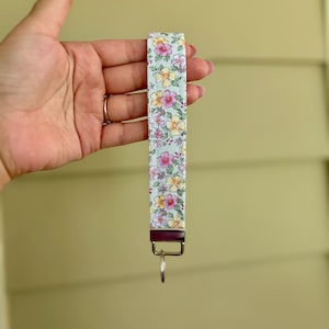 May include: A floral patterned wristlet key fob. The fabric features a light blue background with pink and yellow flowers. The key fob has a silver-colored key ring and a dark metal clasp.