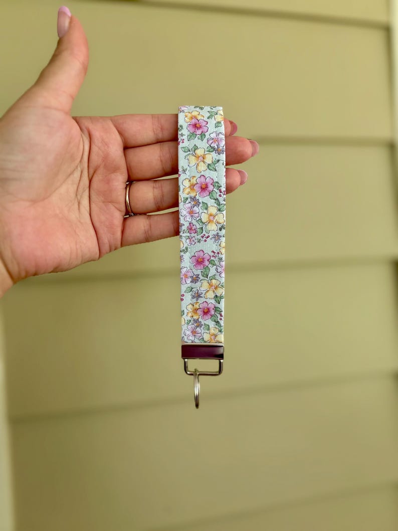 May include: A floral patterned wristlet key fob. The fabric features a light blue background with pink and yellow flowers. The key fob has a silver-colored key ring and a dark metal clasp.