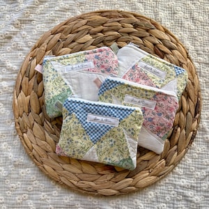 May include: Three quilted zipper pouches featuring a patchwork design of floral and blue gingham fabric patterns. Each pouch has a white rectangular label with the text Loni & Rose stitched onto the front. The pouches are rectangular in shape.