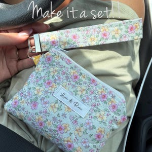 May include: A floral print wristlet and coin purse set. The light green fabric is covered in pink and yellow flowers. The coin purse has a zipper closure and a white label that reads "Luci & Rose". The wristlet has a silver-tone clasp.