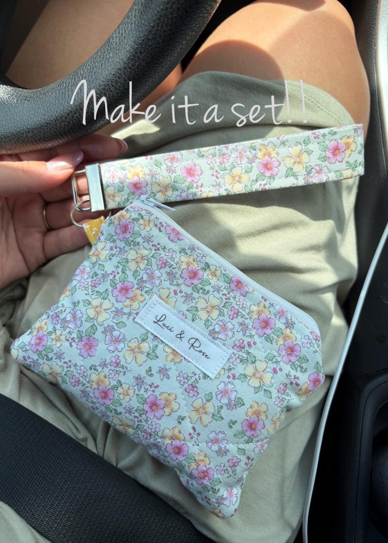 May include: A floral print wristlet and coin purse set. The light green fabric is covered in pink and yellow flowers. The coin purse has a zipper closure and a white label that reads "Luci & Rose". The wristlet has a silver-tone clasp.