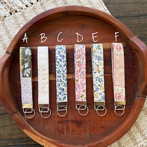 May include: Six fabric key fobs with floral patterns in various colors, including blue, pink, and white. Each fob has a metal ring and is displayed on a wooden tray.