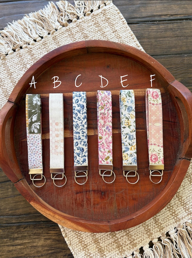 May include: Six fabric key fobs with floral patterns in various colors, including blue, pink, and white. Each fob has a metal ring and is displayed on a wooden tray.