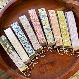 May include: A collection of eight floral patterned wrist keychains in a wooden bowl. The keychains feature various colors, including pink, blue, yellow, and green, with gold-colored hardware. The keychains are arranged in a row, showcasing their different designs.