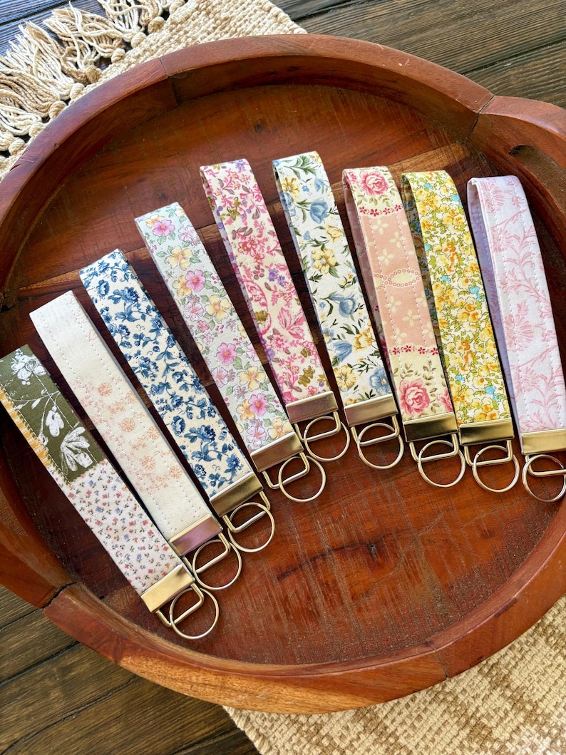 May include: A collection of eight floral patterned wrist keychains in a wooden bowl. The keychains feature various colors, including pink, blue, yellow, and green, with gold-colored hardware. The keychains are arranged in a row, showcasing their different designs.