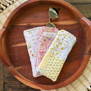 Puede incluir: Tres estuches para gafas con estampado floral en rosa, lavanda y amarillo, con un par de gafas de sol doradas encima. Cada estuche tiene una etiqueta que dice "Lori & Rose". Los estuches se exhiben en una bandeja de madera.