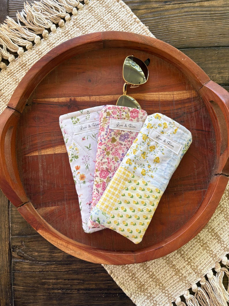 Puede incluir: Tres estuches para gafas con estampado floral en rosa, lavanda y amarillo, con un par de gafas de sol doradas encima. Cada estuche tiene una etiqueta que dice "Lori & Rose". Los estuches se exhiben en una bandeja de madera.