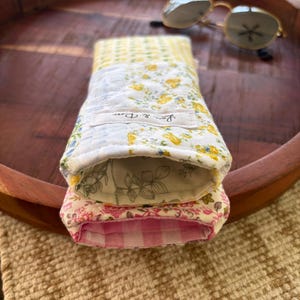 Puede incluir: Dos estuches de tela para gafas, uno encima del otro, con estampados florales y de cuadros. El estuche superior tiene flores amarillas sobre fondo blanco, y el inferior flores rosas. Gafas de sol sobre una bandeja de madera.