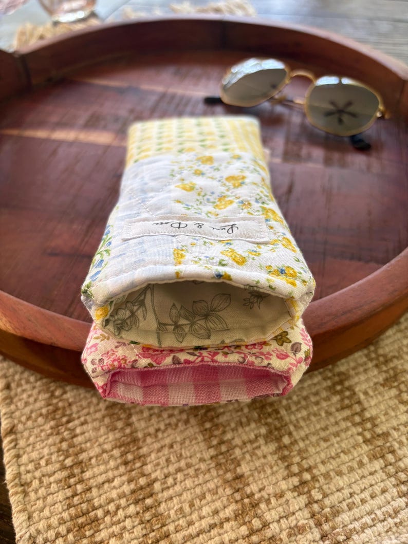Puede incluir: Dos estuches de tela para gafas, uno encima del otro, con estampados florales y de cuadros. El estuche superior tiene flores amarillas sobre fondo blanco, y el inferior flores rosas. Gafas de sol sobre una bandeja de madera.