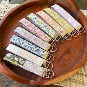 May include: A collection of key fobs with floral patterns in various colors, including pink, blue, yellow, and green. Each fob has a gold-colored metal ring and a fabric strap. The key fobs are arranged in a wooden bowl.