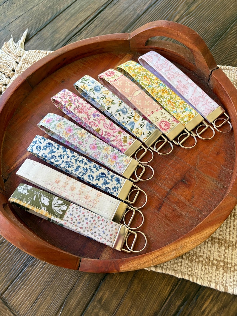 May include: A collection of key fobs with floral patterns in various colors, including pink, blue, yellow, and green. Each fob has a gold-colored metal ring and a fabric strap. The key fobs are arranged in a wooden bowl.