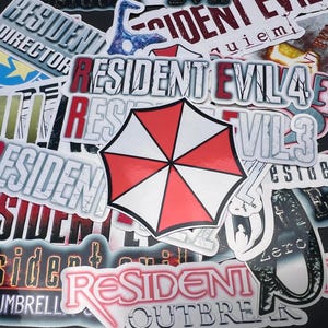 May include: A selection of stickers showcasing the "Resident Evil" franchise, including the Umbrella Corporation logo and game titles. The stickers feature the red and white umbrella emblem and text in bold fonts. The stickers are in various sizes and shapes.