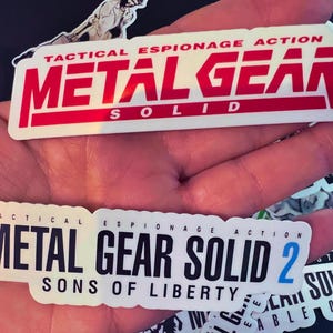 May include: Two white stickers with black text. The top sticker reads "Tactical Espionage Action Metal Gear Solid". The bottom sticker reads "Tactical Espionage Action Metal Gear Solid 2 Sons of Liberty".