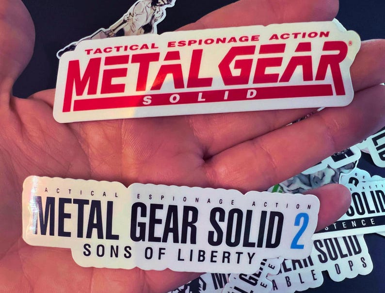 May include: Two white stickers with black text. The top sticker reads "Tactical Espionage Action Metal Gear Solid". The bottom sticker reads "Tactical Espionage Action Metal Gear Solid 2 Sons of Liberty".