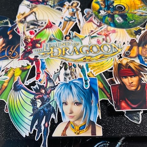 May include: A selection of vibrant stickers displaying characters and the title logo from the video game "The Legend of Dragoon". The stickers depict diverse characters with detailed artwork, including a woman with blue hair and a man with a red headband.