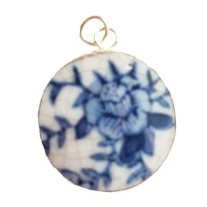 May include: A circular pendant featuring a white ceramic base with a blue floral pattern and a distinct crackle glaze finish. The pendant is topped with a small, thin metal loop for attachment, showcasing a classic blue and white color scheme.