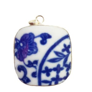 May include: A square-shaped pendant featuring a white ceramic base with a blue floral and vine pattern. The pendant has rounded corners and a small gold-toned metal loop at the top for attachment, displaying a classic blue and white porcelain design.